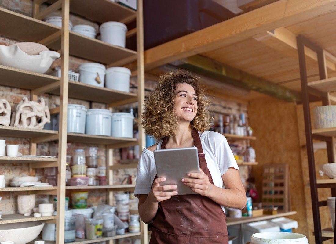 Business Insurance - Portrait of a Cheerful Young Small Business Owner Holding a Tablet in her Hands as she Stands Inside her Pottery and Crafts Studio