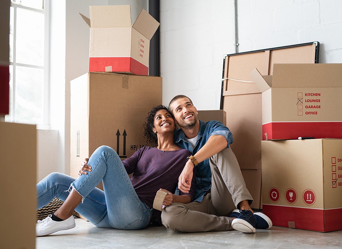 Insurance Solutions - View of a Happy Young Married Couple Sitting Next to Moving Boxes on the Floor of Their New Apartment