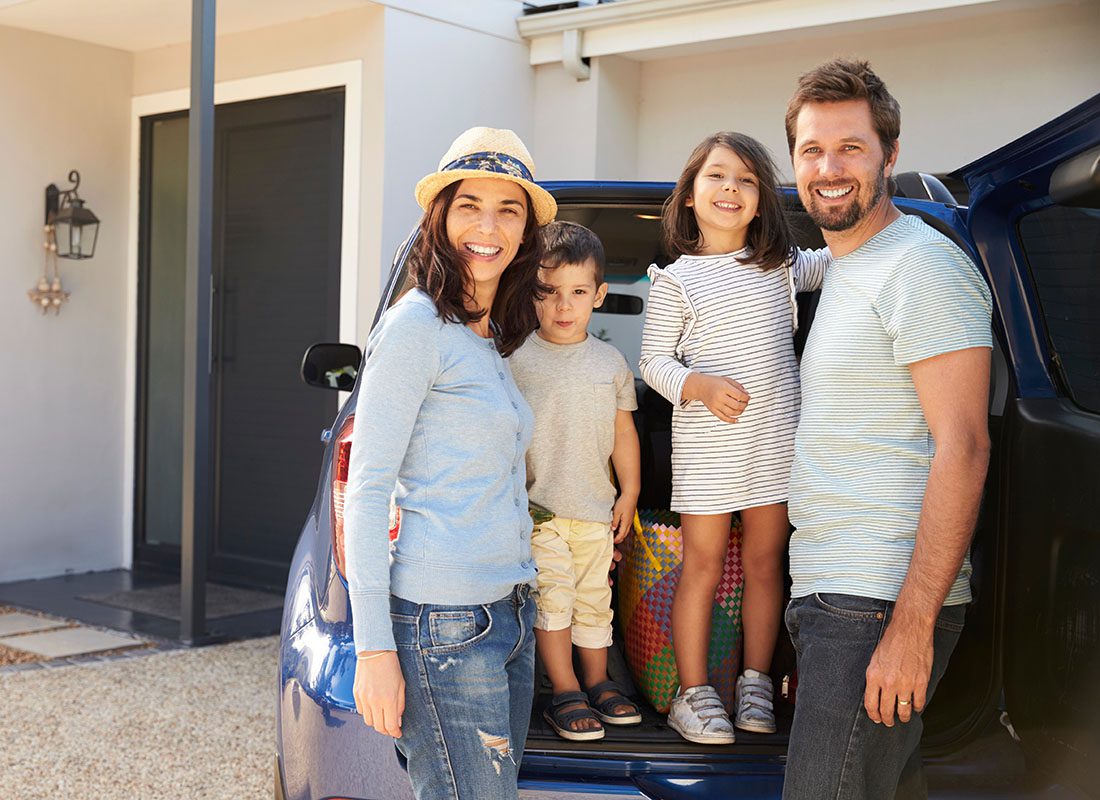 Personal Insurance - Portrait of a Cheerful Family with Two Young Kids Standing Next to Their SUV Parked Next to the House Ready to Go on a Summer Vacation