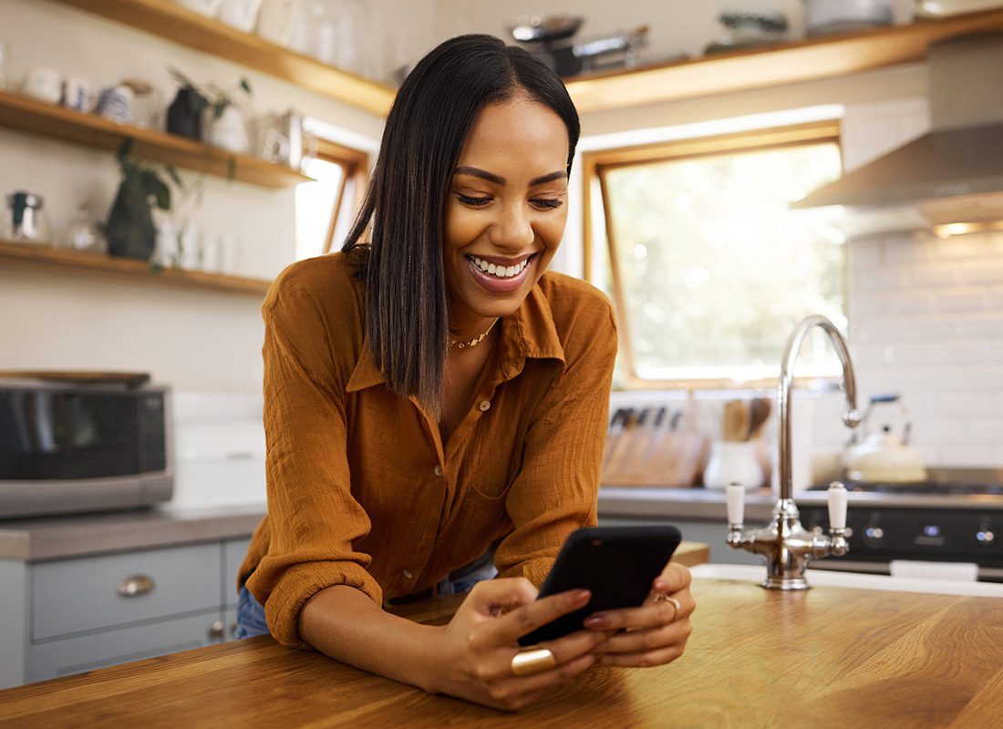 Read Our Reviews - Closeup Portrait of a Cheerful Young Woman Leaning Against the Wooden Countertop in her Kitchen While She Uses a Phone